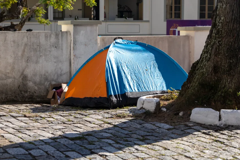 A orange-and-blue tent set up next to a tree in a paved courtyard and a cardboard box of posessions next to the tent entrance.
